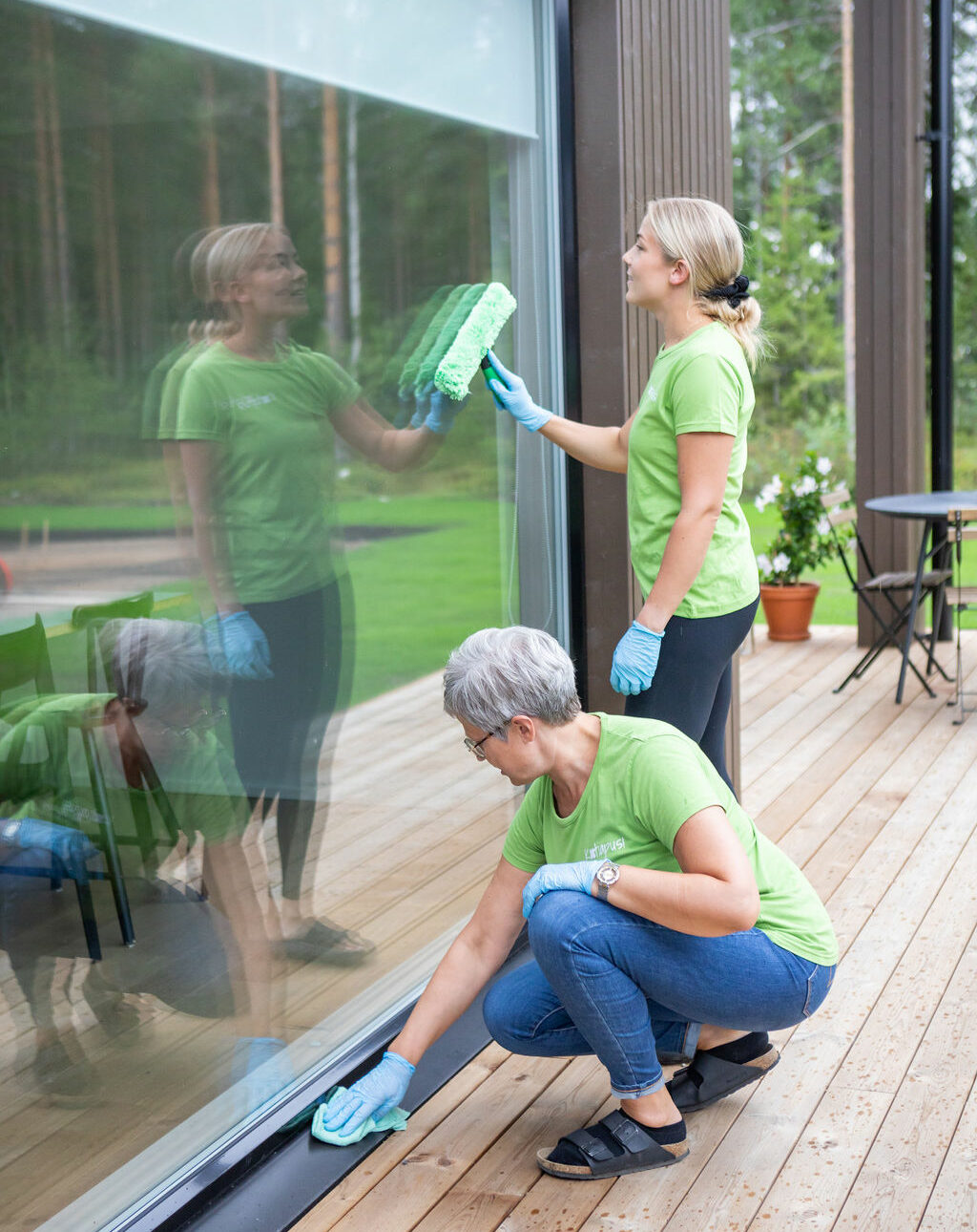 Kaksi vihreisiin siivouspalvelun työpaitoihin pukeutunutta henkilöä pesemässä huolella modernin asunnon suurta ikkunaa ulkopuolelta.