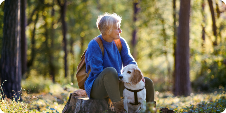 Iäkäs nainen koiran kanssa ulkoilemassa metsässä. Nainen istuu kannolla.