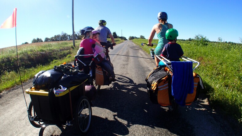 Perhe pyöräilemässä maantiellä pellon keskellä. Pyörissä on mukana peräkärryt.